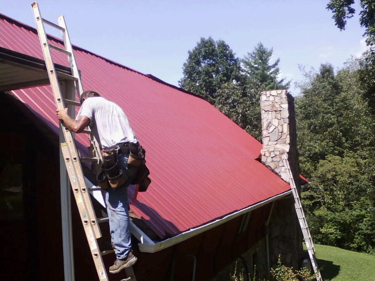 Roofer on a ladder beside a red metal roof and stone chimney — Soffit & Fascia Repair by Roofing Brothers in Buffalo Valley